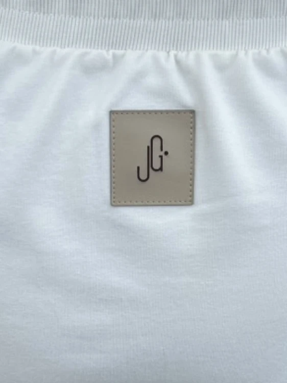 Dekoracja długiej spódnicy z rozcięciem w kolorze jasnego ecru umieszczona z tyłu pod pasem. Naszywka z logo JG