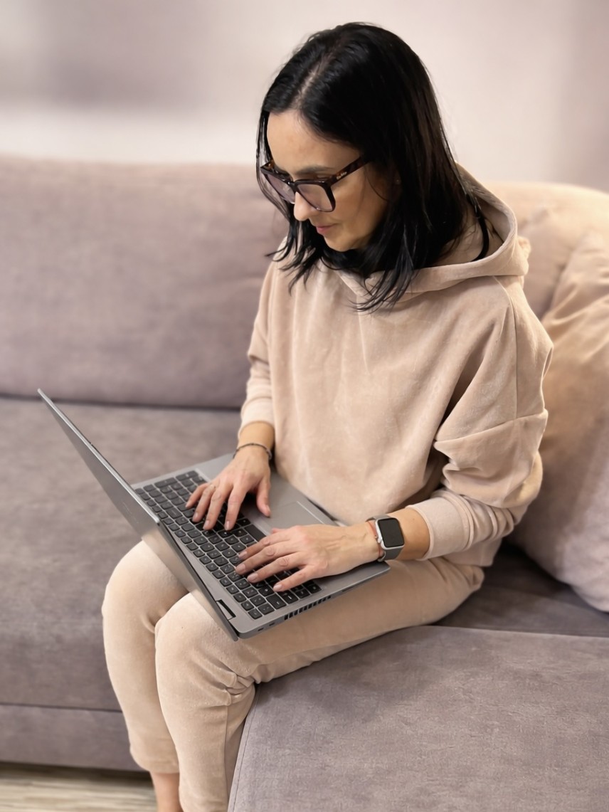 Grafika prezentuje kobietę siedzącą na kanapie . Kobieta trzyma na kolanach  laptop odpowiadając na pytania z sekcji FAQ